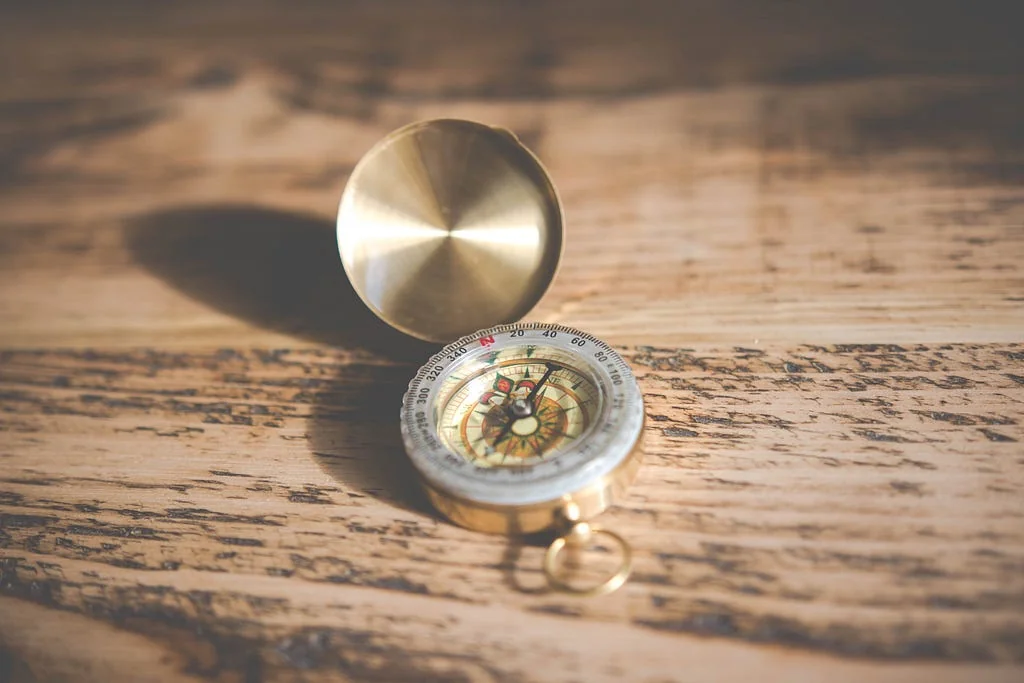 A brass pocket compass lying open on a weathered wooden table, its lid raised and its needle pointing off to one side.
