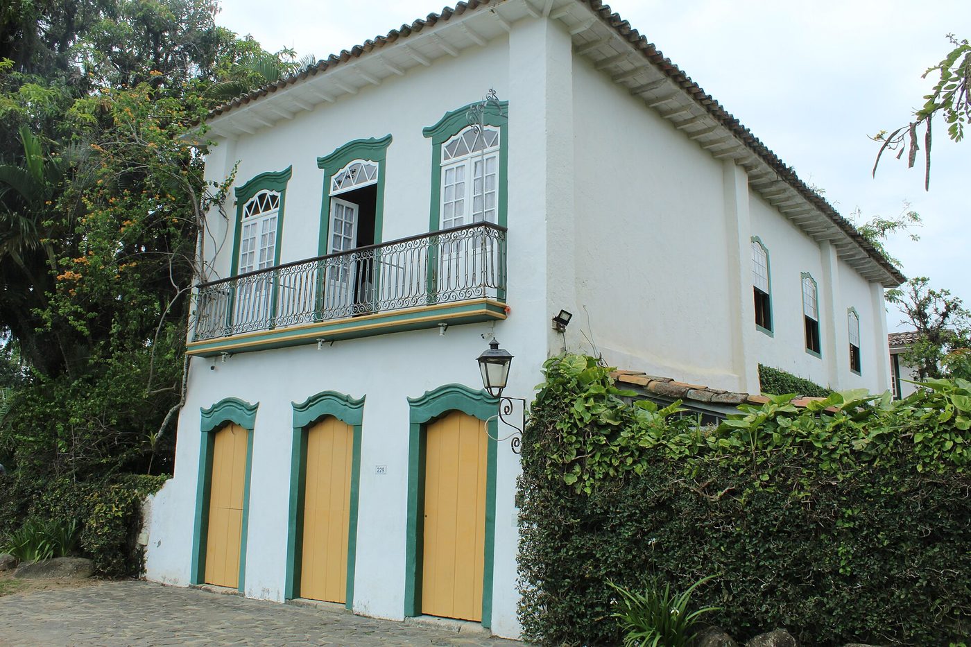 A two-story colonial townhouse (casarão) in Paraty with white walls, green-trimmed windows and doors, yellow door panels, a wrought-iron upper balcony, and a terracotta roof, the number 229 beside a doorway.