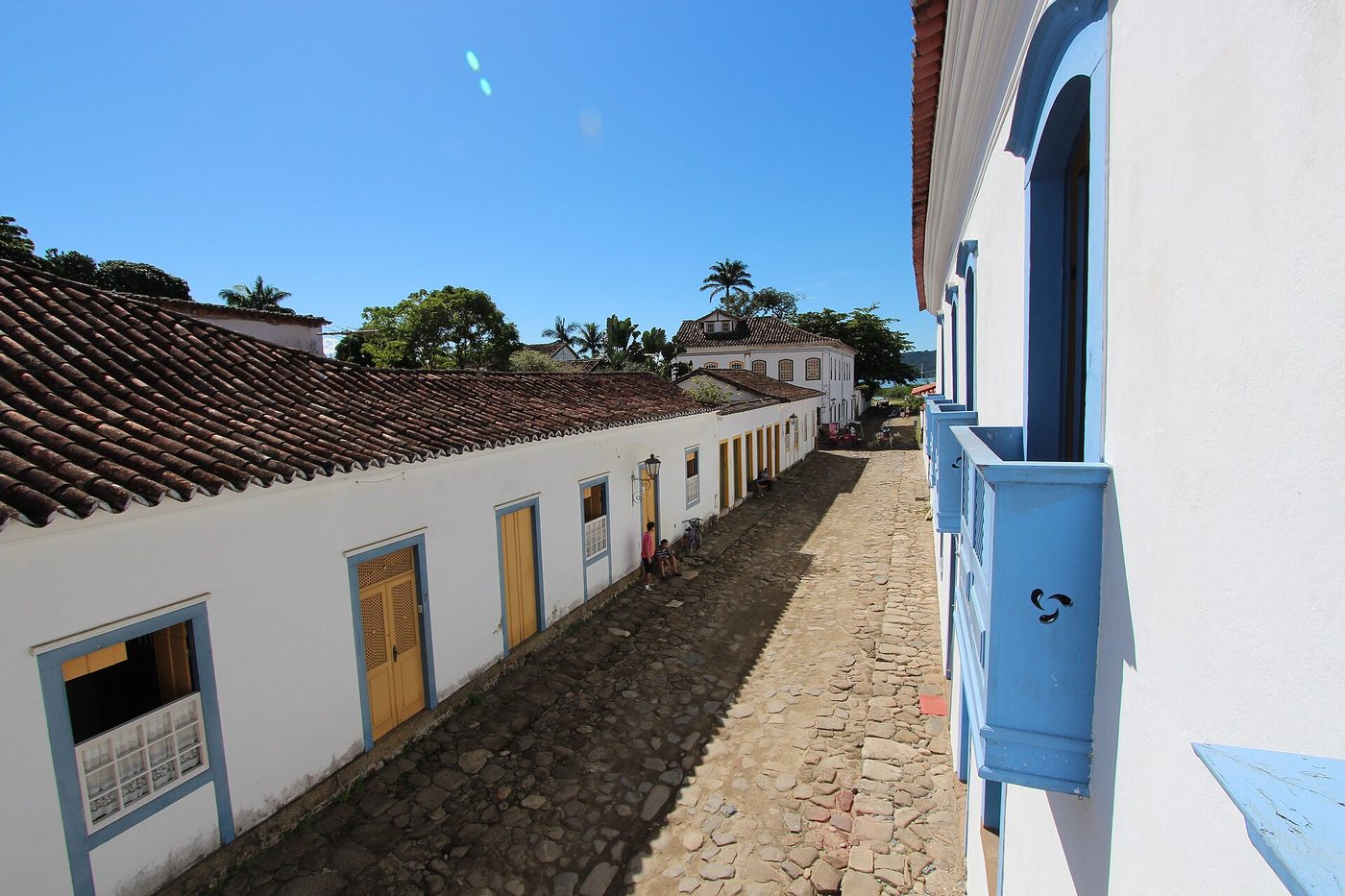 Looking down a Paraty street from rooftop height: whitewashed single-story colonial houses with blue and yellow window frames, uneven cobblestones, terracotta-tiled roofs, and the bay visible at the far end.