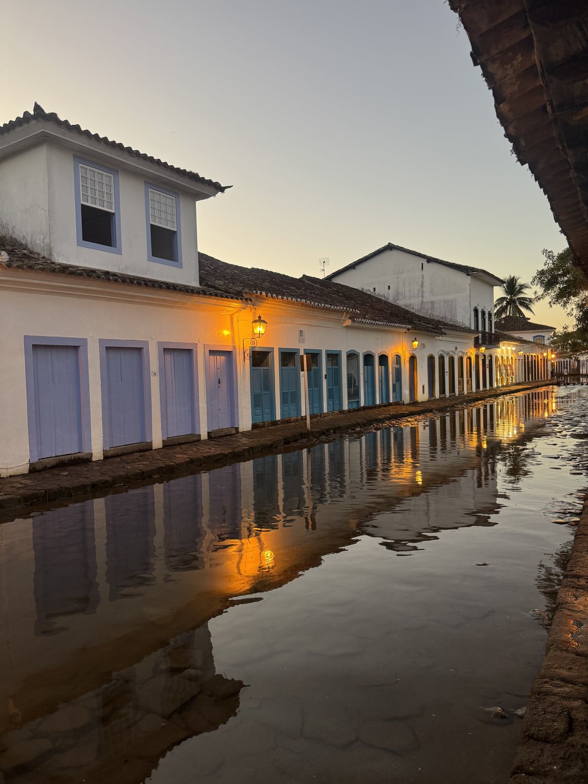 Dusk in Paraty during a spring tide: seawater covers the cobbled street and mirrors a row of lantern-lit whitewashed houses with blue and lavender doors, a church tower rising behind them.