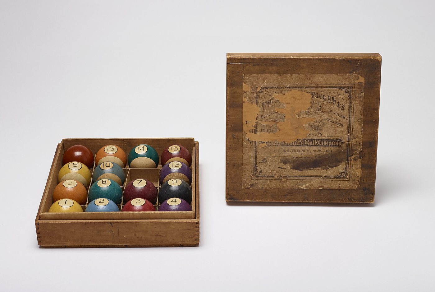 A wooden box of antique numbered billiard balls in muted reds, greens, blues and yellows, with its worn lid propped beside it.