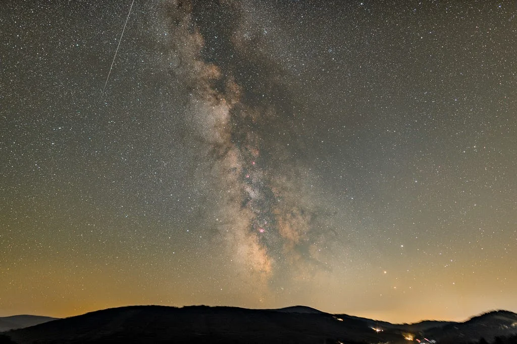 The Milky Way arching over a dark ridge of hills at night, a meteor streak in the upper left and a faint glow of town lights along the horizon.