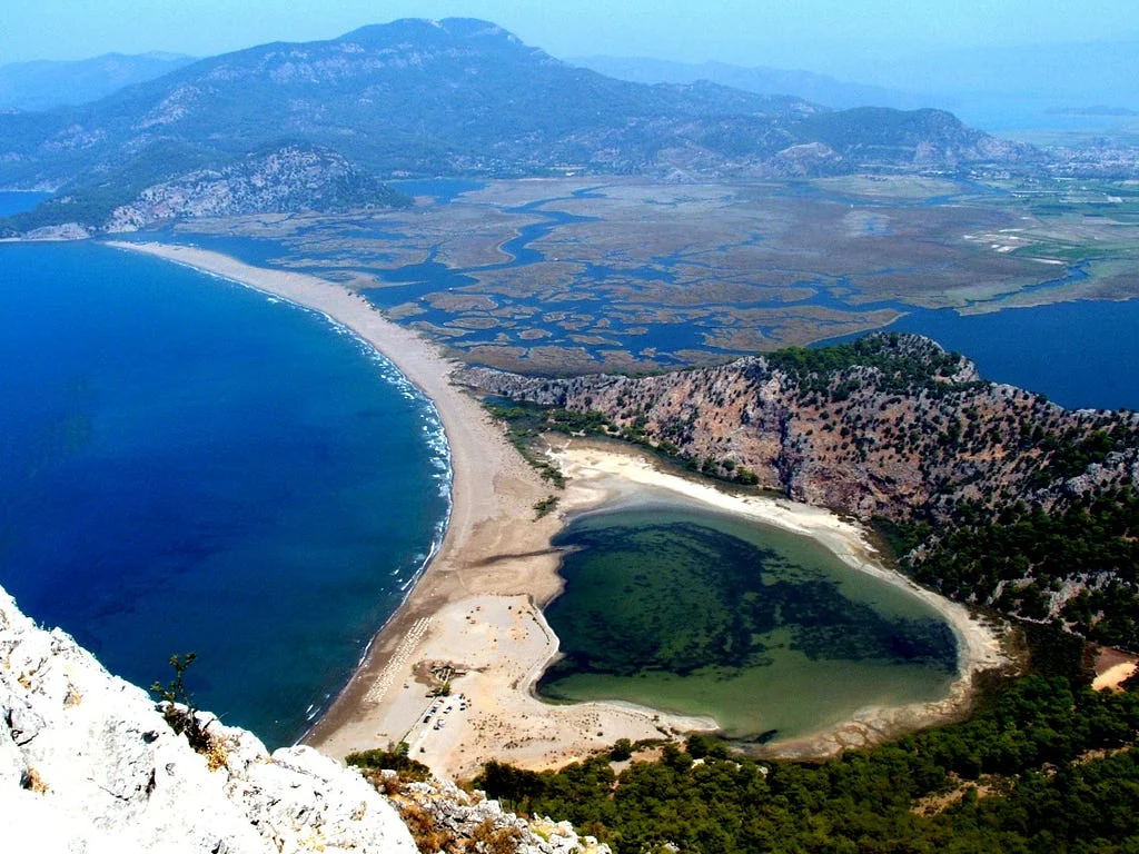 Iztuzu beach from a clifftop: a long curved sand spit dividing the deep blue Mediterranean from the reed-lined channels of the Dalyan delta, with a green lagoon at the near end and mountains across the water.